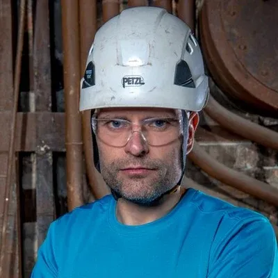 Koolitaja Toomas Holmberg valges kaitsekiivris ja kaitseprillides, taustal tööstushoone torustik